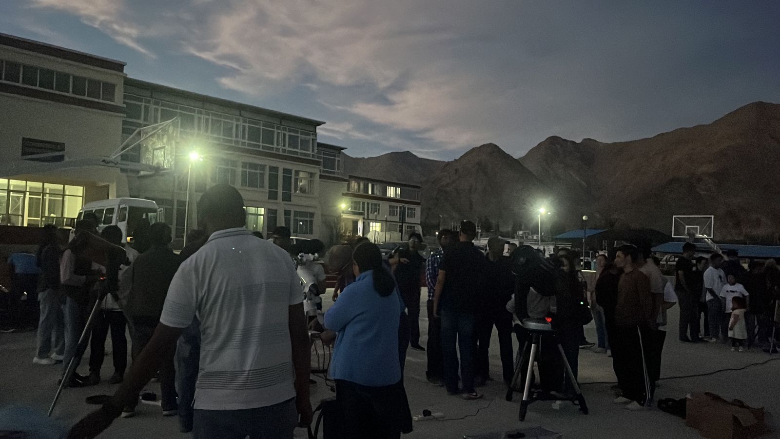 A crowd of people stands outside at night, with mountains illuminated in the background under a starry sky.